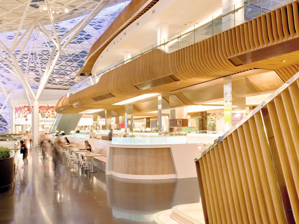 Modernes Einkaufszentrum mit Glasdach, geschwungenen Holzelementen und Menschen, die an Tischen sitzen.