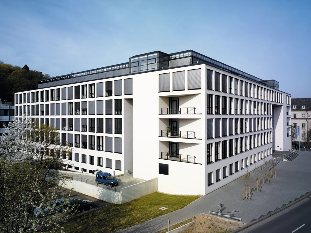 Modernes weißes Gebäude mit vielen Fenstern, Balkonen und blühenden Bäumen im Vordergrund.