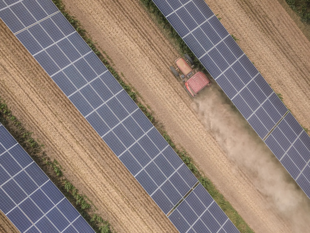 Ein Traktor pflügt ein Feld zwischen Reihen von Solarpanels aus der Vogelperspektive.