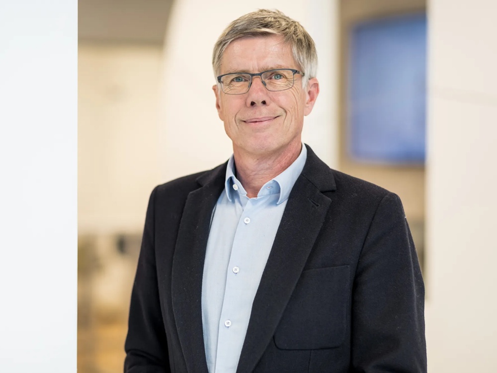 Ein Mann mit Brille in Anzug und Hemd steht lächelnd in einem hellen Raum.