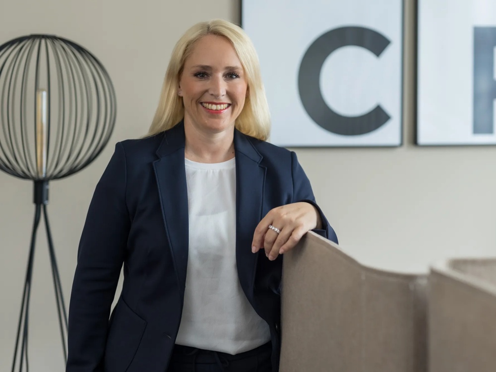 A smiling woman in a dark blazer standing next to a lampshade, with large letters in the background.