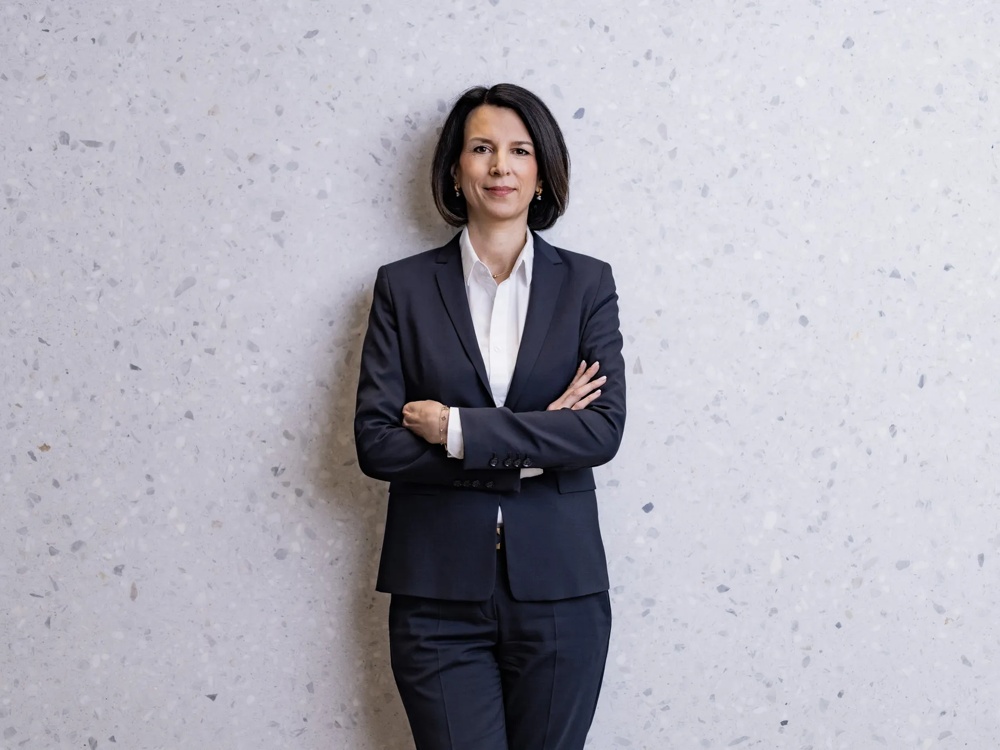 Eine Person in einem dunklen Anzug steht mit verschränkten Armen vor einer hellgrauen, gesprenkelten Wand.