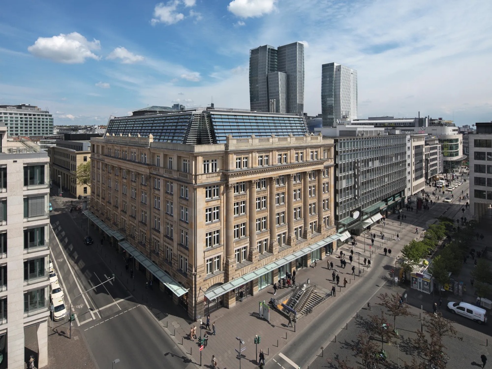 Großes, historisches Eckgebäude mit Glasdach, umgeben von Straßen und Fußgängern bei Sonnenschein.