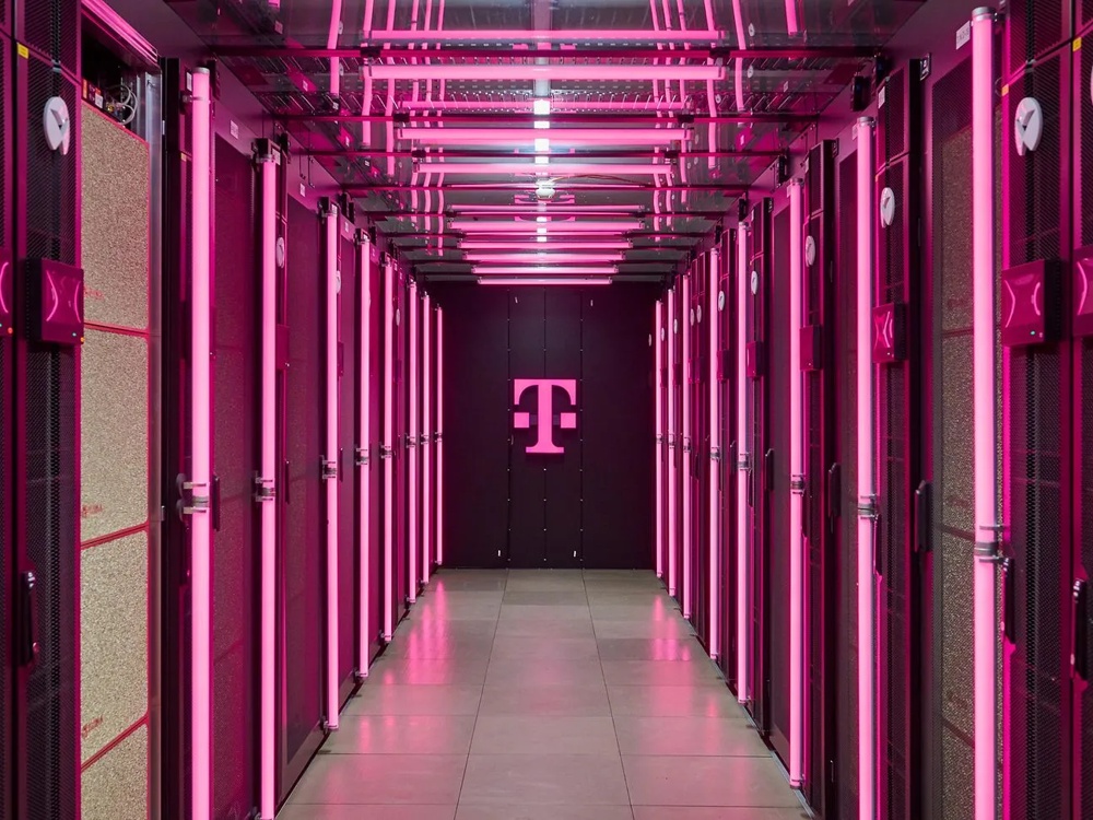 View through a magenta-lit Deutsche Telekom server room. Tall server racks line the corridor, with the Telekom T logo at the far end.