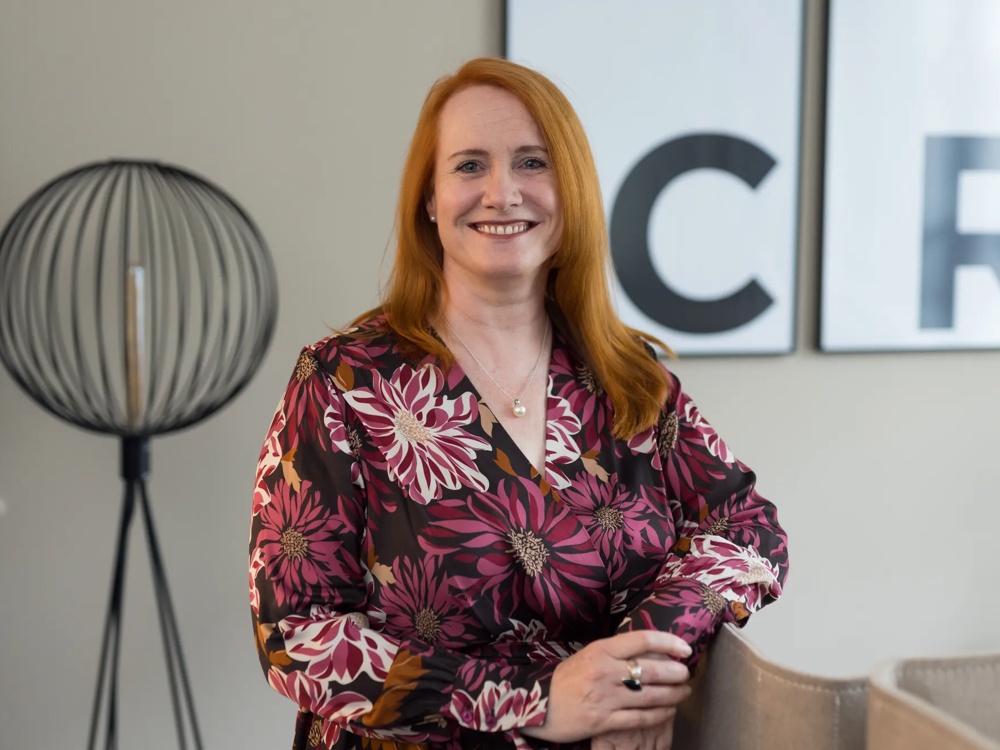 Woman with red hair and floral dress smiling in front of a floor lamp and two letter pictures.