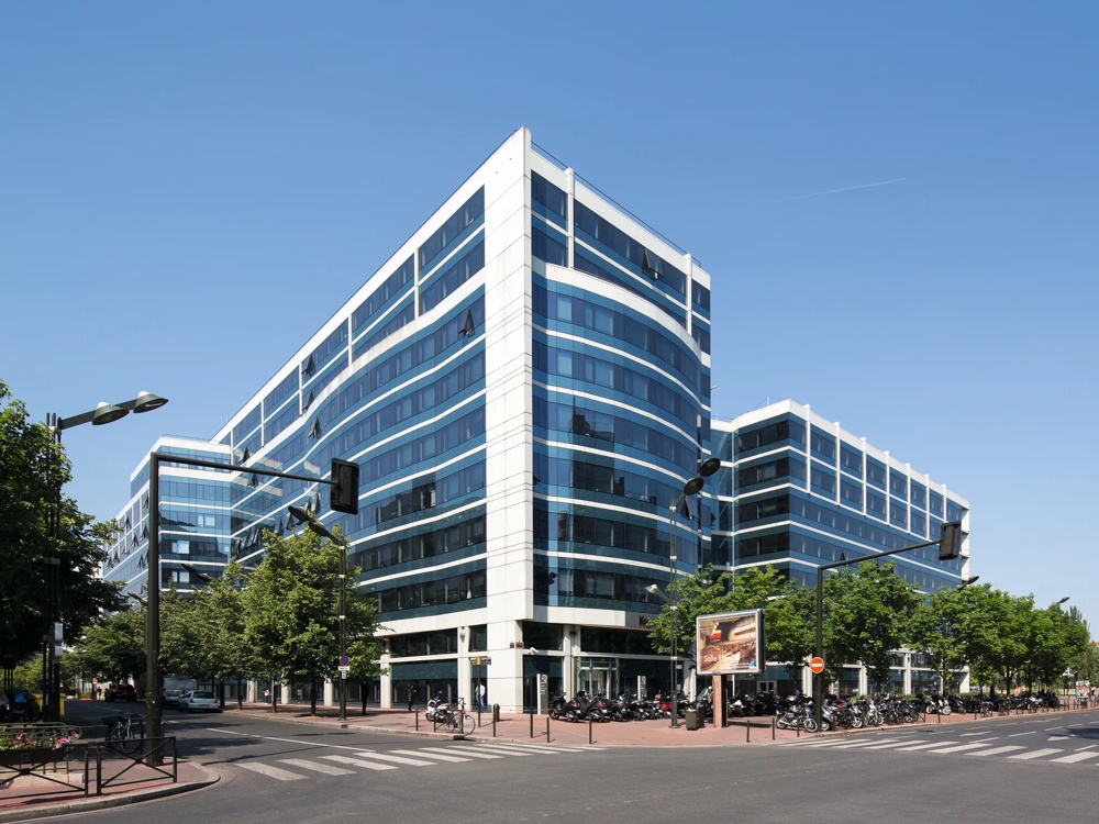 Modernes Bürogebäude mit Glasfassade an einer Straßenecke, umgeben von Bäumen und geparkten Fahrrädern.