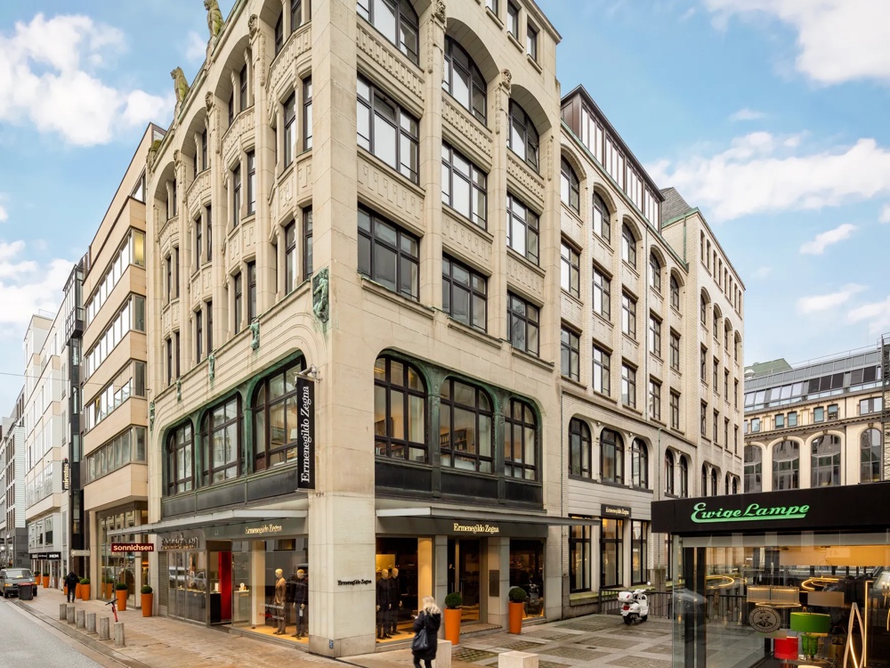 Eckgebäude mit großen Fenstern und dekorativen Details, Geschäfte im Erdgeschoss, blauer Himmel im Hintergrund.