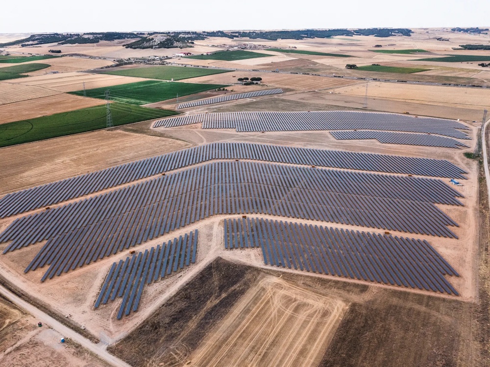 Große Solaranlage auf weitläufigen Feldern, umgeben von landwirtschaftlichen Flächen und Hügeln im Hintergrund.