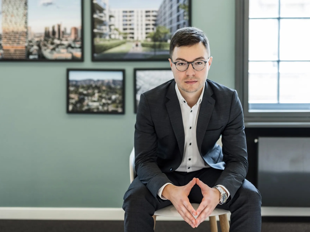 A man in a suit sitting in front of a grey wall with framed pictures.