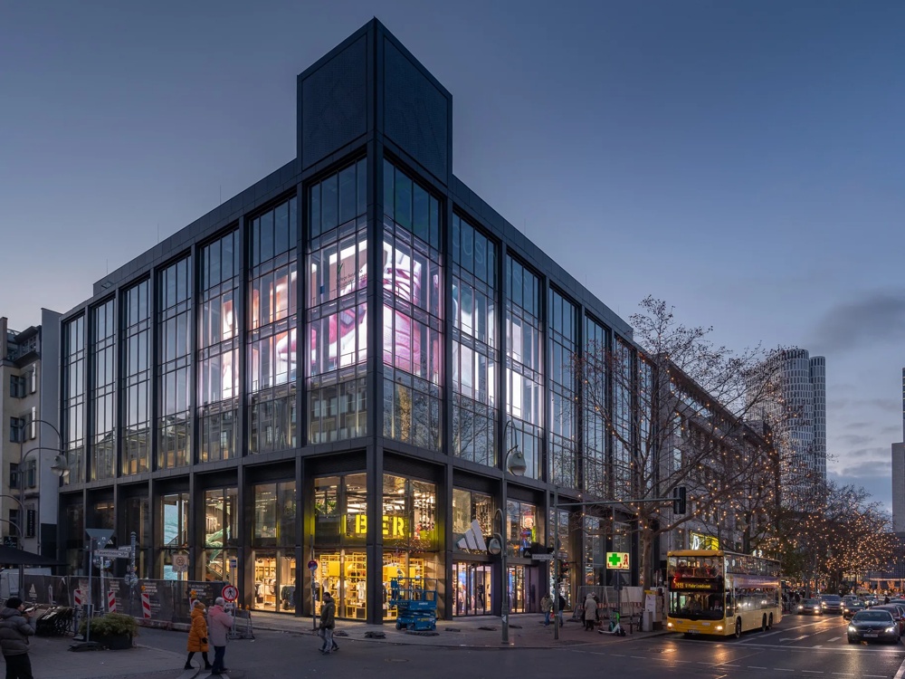 Modernes Glasgebäude bei Dämmerung, leuchtende Werbeanzeigen, Menschen und Bus auf belebter Straße.
