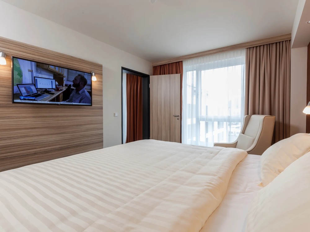 Modernes Hotelzimmer mit Doppelbett, Fernseher, Sessel und großem Fenster mit Vorhängen.