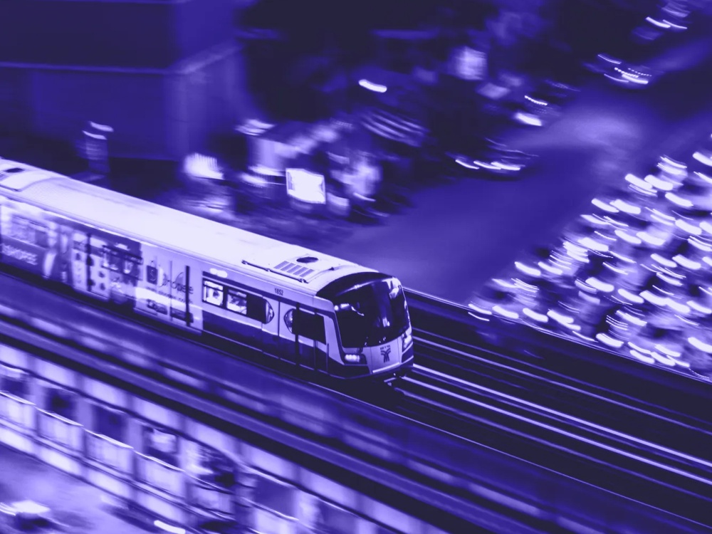 Ein Zug fährt nachts auf erhöhten Gleisen, umgeben von verschwommenen Lichtern und Gebäuden in Blautönen.