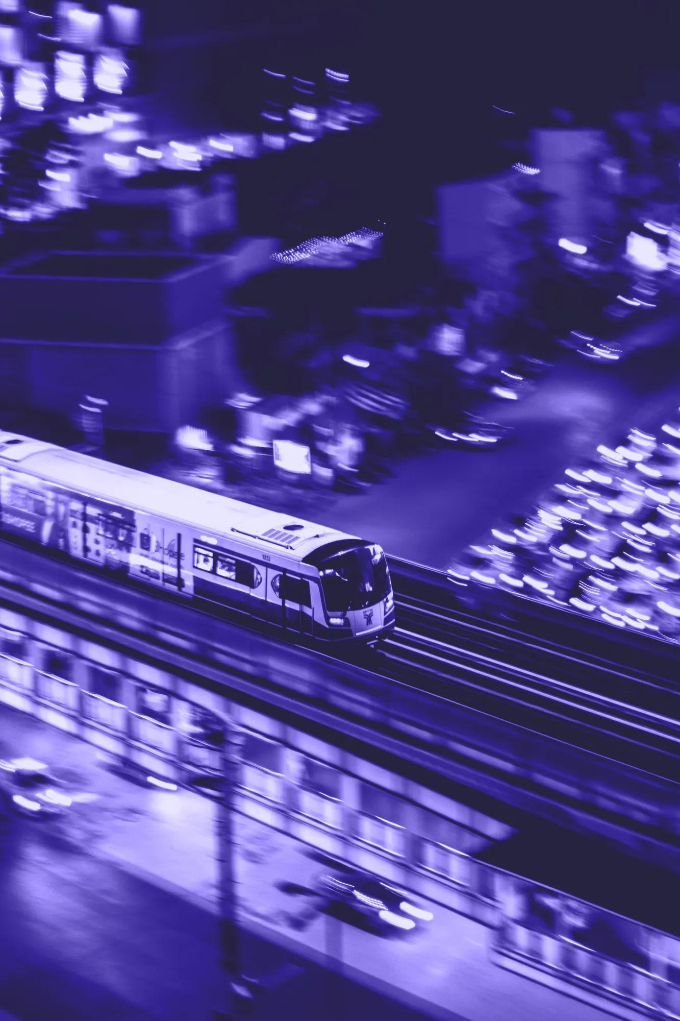 Ein Zug fährt nachts auf erhöhten Gleisen, umgeben von verschwommenen Lichtern und Gebäuden in Blautönen.