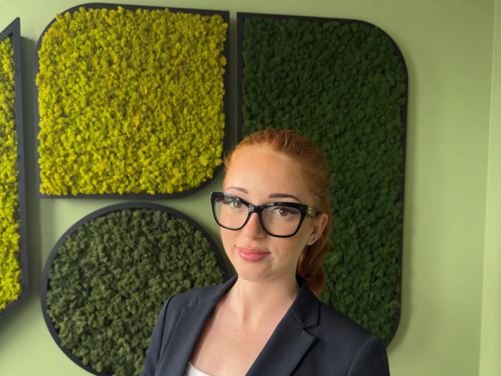 Junge Frau mit markanter schwarzer Brille und einem Sakko über weißem Shirt. Im Hintergrund eine grüne Mosswand.