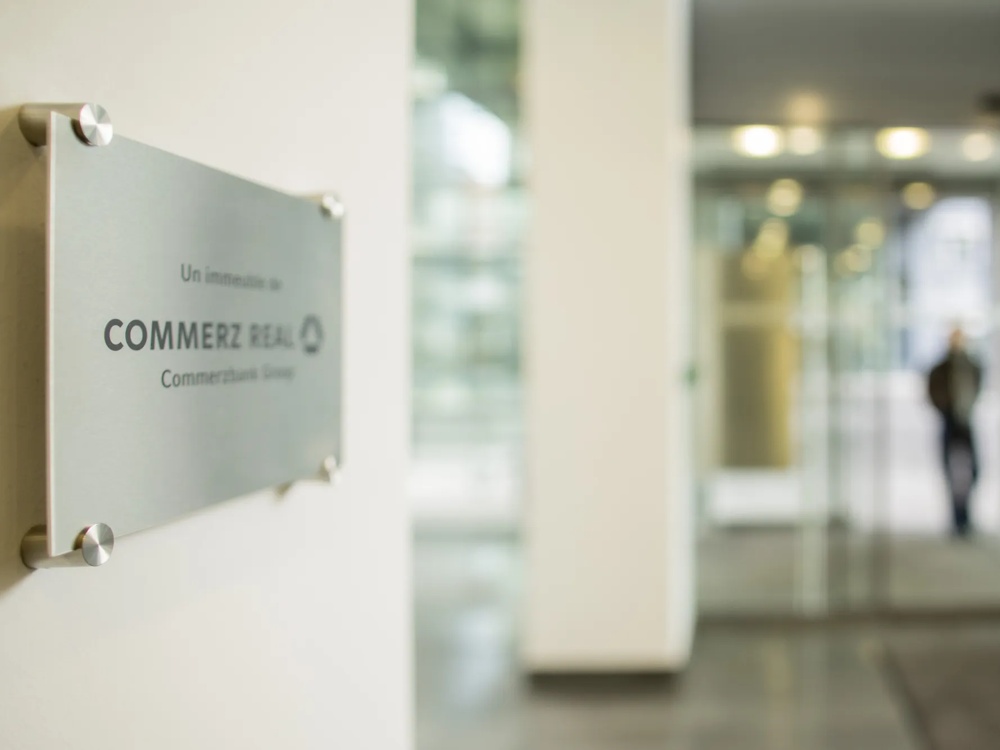 Ein Schild mit der Aufschrift "Commerz Real" hängt an einer Wand in einem modernen, hellen Gebäude.