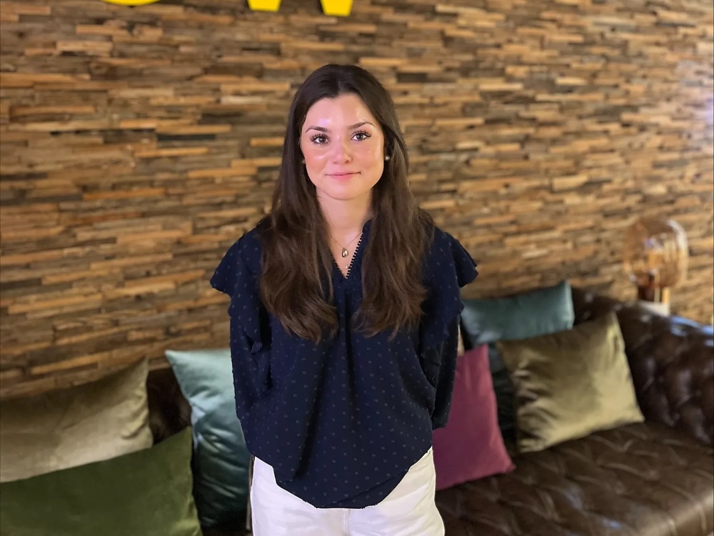Junge Frau mit langen brünetten Haaren und schwarzer Bluse und weißer Hose vor einer Holzwand mit den Schriftzug Digitalwerk