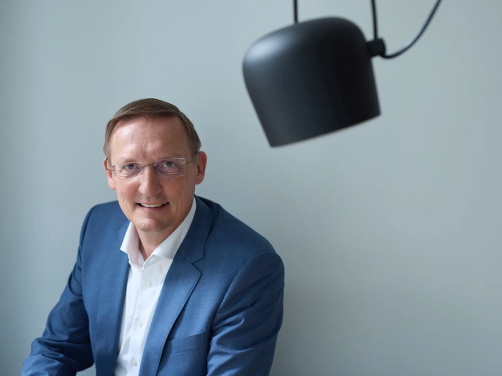 Commerz Real employee Ulrich Pflügler, Senior Key Account Manager for Sales Private and Business Clients SW, is shown. He is looking at the camera, wearing a light blue blazer and white shirt, sitting under a modern black lamp.