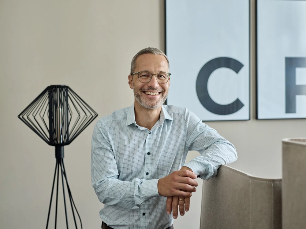 A smiling man in a shirt and glasses leaning on an armchair, behind him a lamp and two pictures with letters.