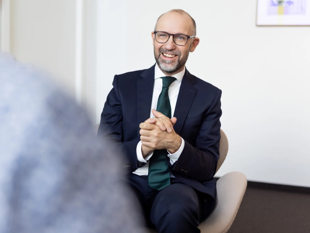 Ein lächelnder Mann in Anzug und Krawatte sitzt auf einem Stuhl in einem Büro.