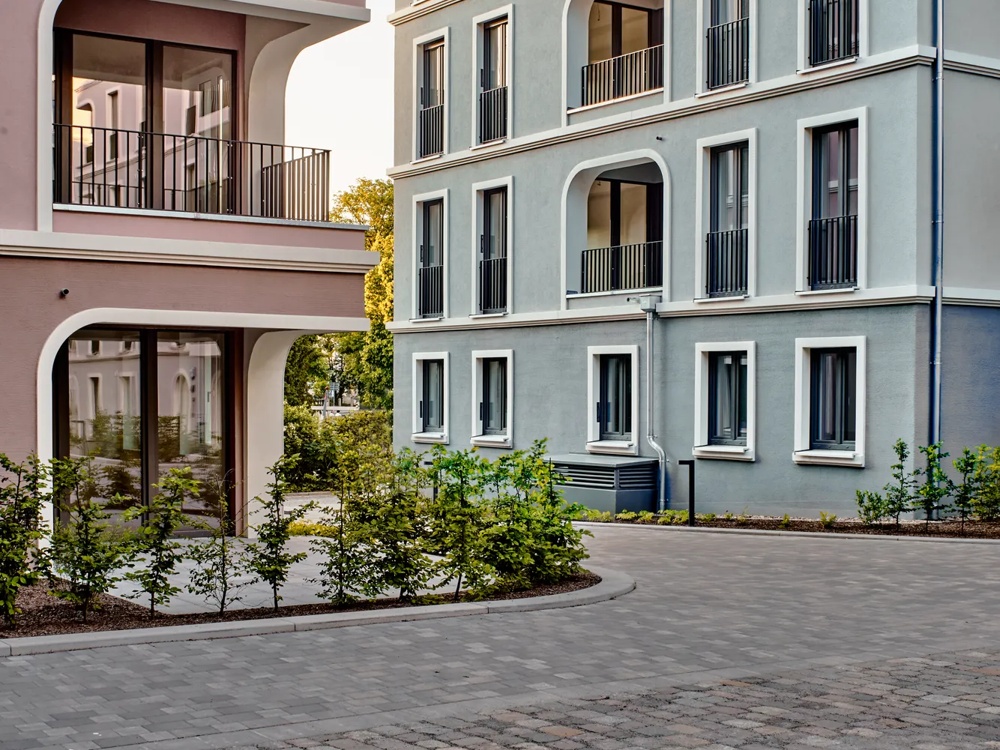 Two modern residential buildings with balconies, one pink and the other grey, at sunset.