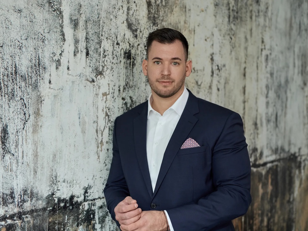 A man is wearing a black suit without a tie and a pocket square. He is standing in front of a marbled wall.