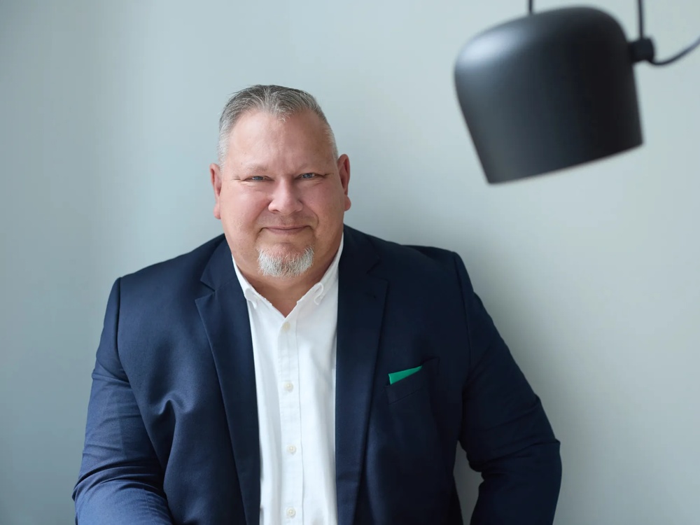 Commerz Real employee Claus André Krüger, Senior Key Account Manager for Sales Private and Business Clients SW, is presented. He is sitting in front of a light grey wall with a black ceiling light, wearing a dark blue jacket and looking calmly into the camera.