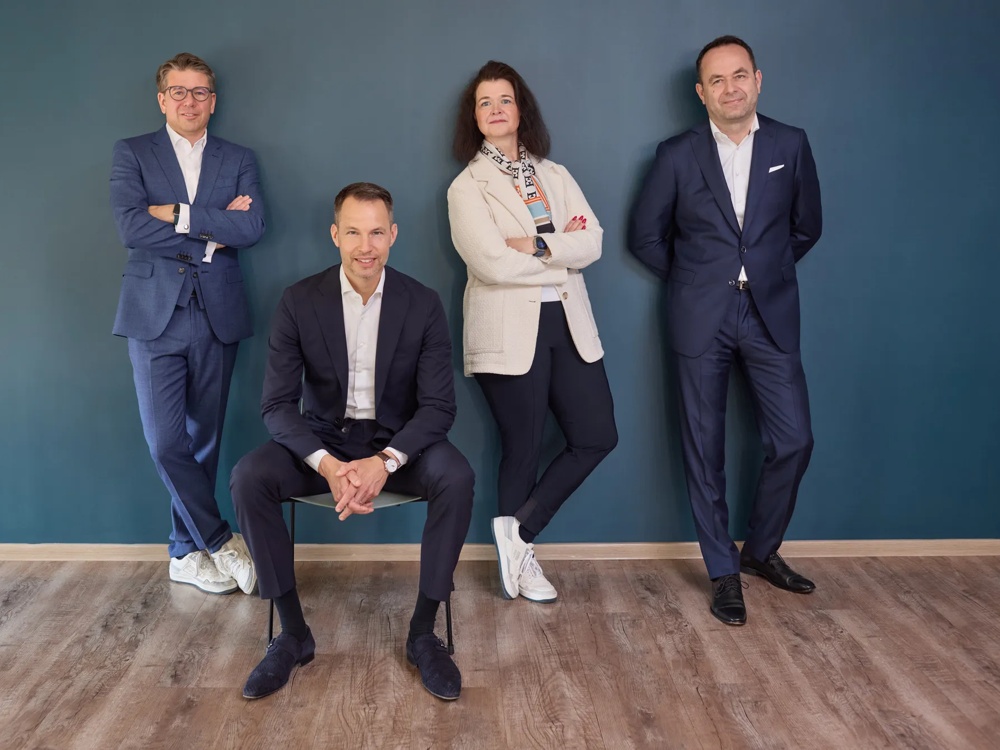 Vier Personen in Anzügen vor einer blauen Wand, drei stehen, einer sitzt auf einem Stuhl.