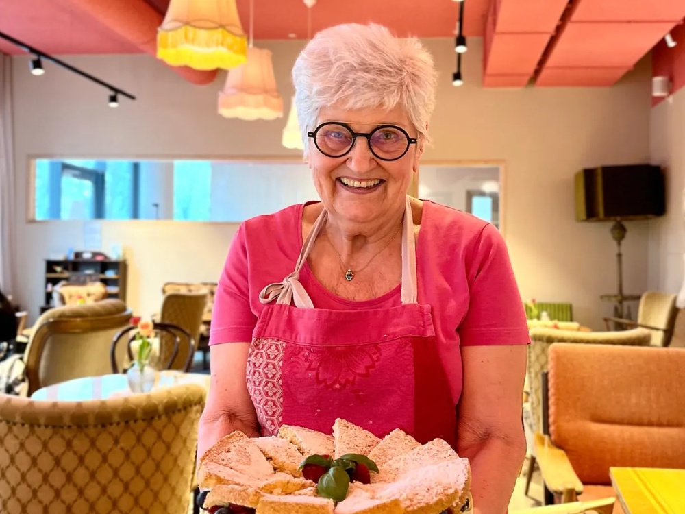 Ältere Frau in rosa Schürze hält eine Sahnetorte mit Beeren in einem gemütlichen Café.