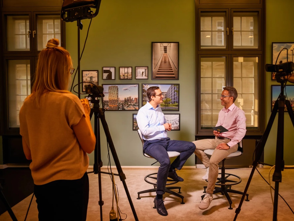 Zwei Männer sitzen auf Hockern in einem Interview, während eine Frau sie filmt; Bilder an grüner Wand im Hintergrund.