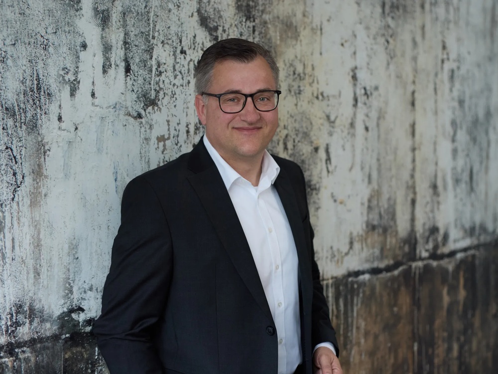 Commerz Real employee Jens Burkhard, Senior Key Account Manager for Sales Private and Business Clients NOM, is represented. He is standing in front of a rough, textured wall, wearing a dark suit with a white shirt and smiling lightly.