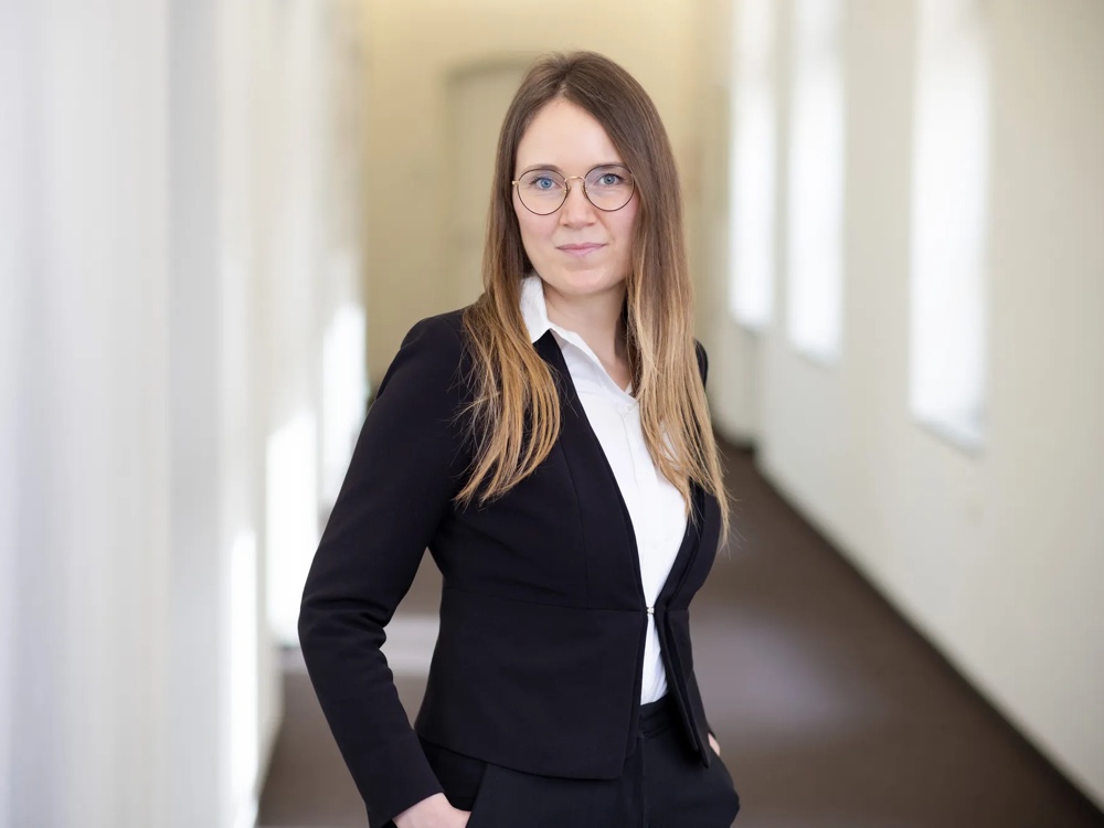 A person in a suit stands in a bright corridor, looking friendly into the camera.