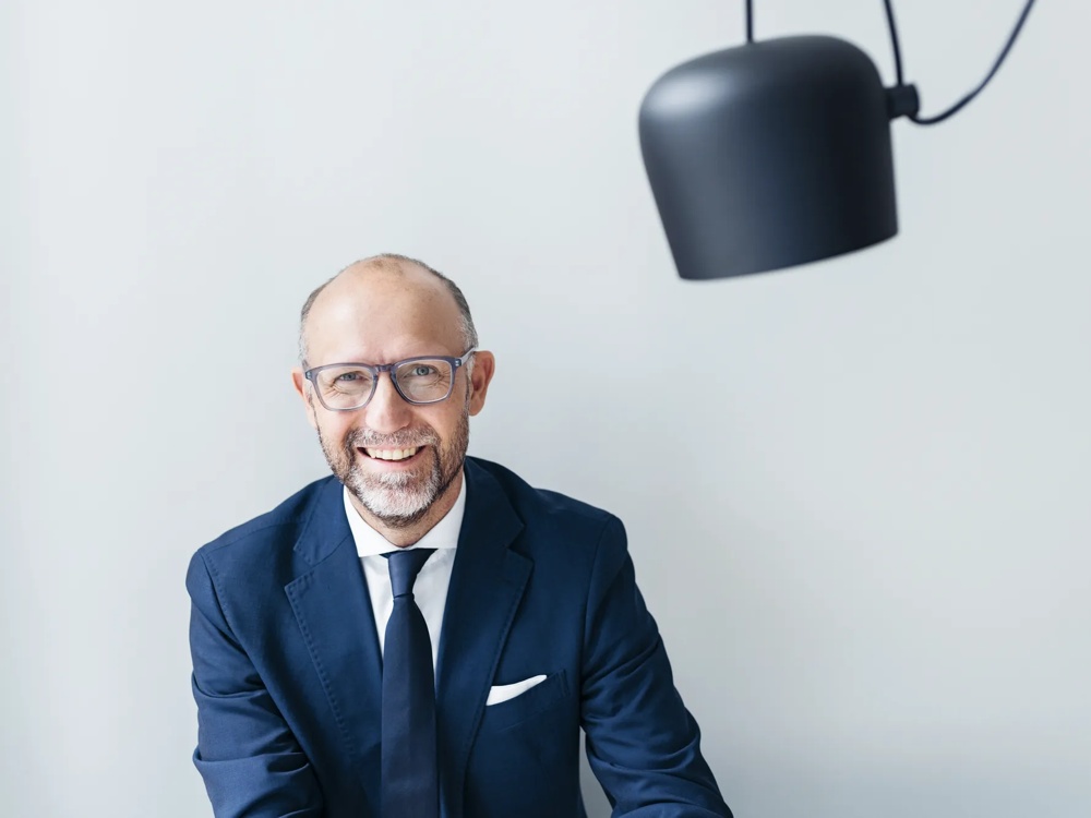 Ein lächelnder Mann im Anzug sitzt unter einer schwarzen Hängelampe vor hellem Hintergrund.