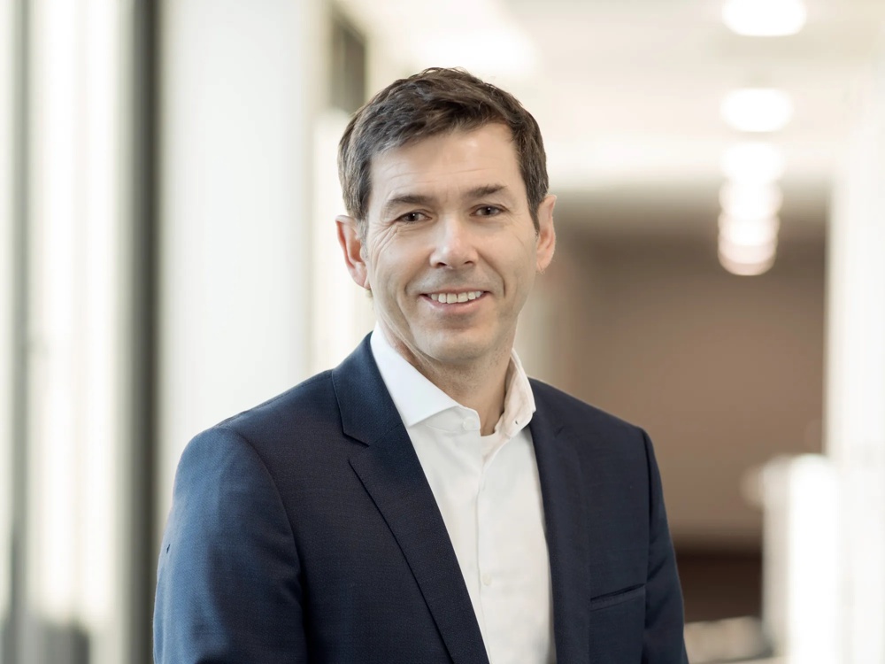 A man in a dark blue suit smiling in a bright hallway.
