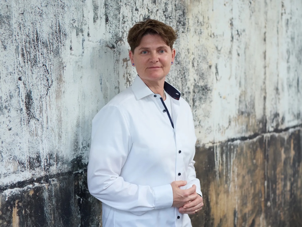 Person in weißem Hemd lehnt an einer verwitterten, strukturierten Wand und schaut in die Kamera.