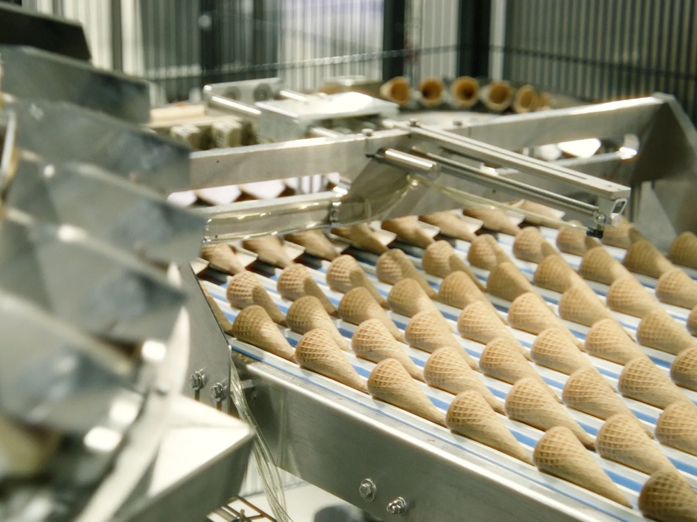 Zwei Fabrikationsmitarbeiter stehen in weißen Kitteln gebeugt über einer Eiswaffelproduktionsstraße und begutachten die Reihen der frischgebackenen Hörnchen.