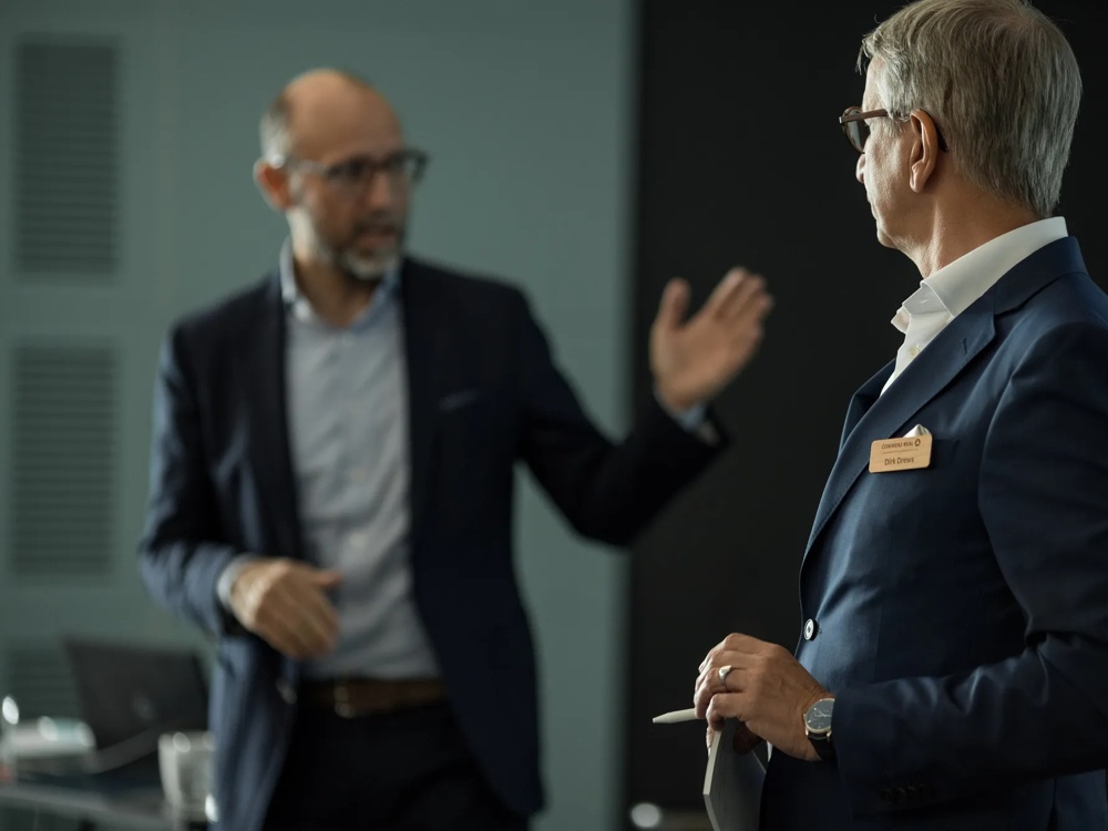 Zwei Männer in Anzügen diskutieren in einem Büro, einer hält Papiere, der andere gestikuliert.