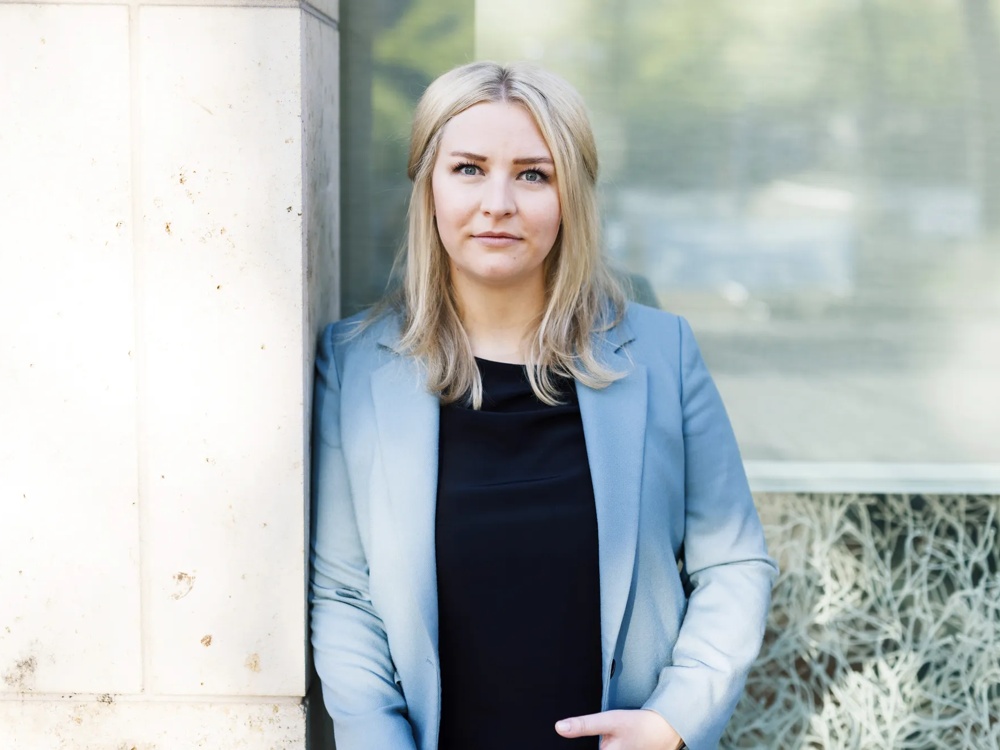 A woman in a blue blazer standing in front of a window looking into the camera.