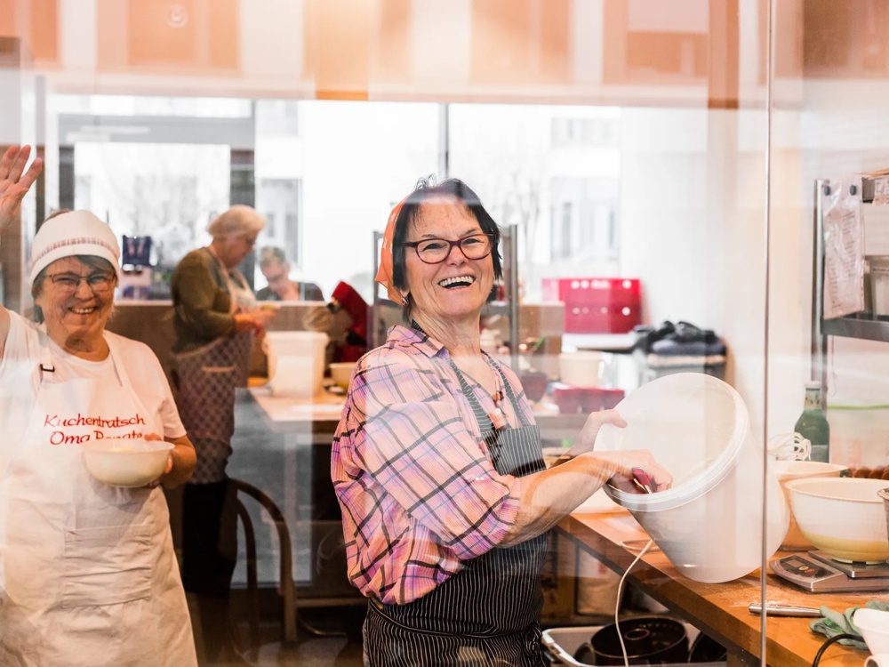 Zwei lachende Frauen in einer Bäckerei, eine winkt, die andere rührt Teig in einer Schüssel.