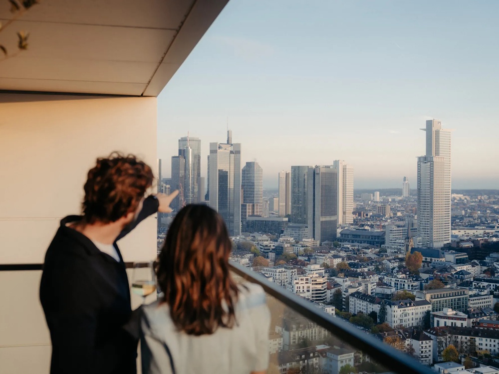 Zwei Personen auf Balkon einer modernen Wohnung mit Blick über die Stadt.