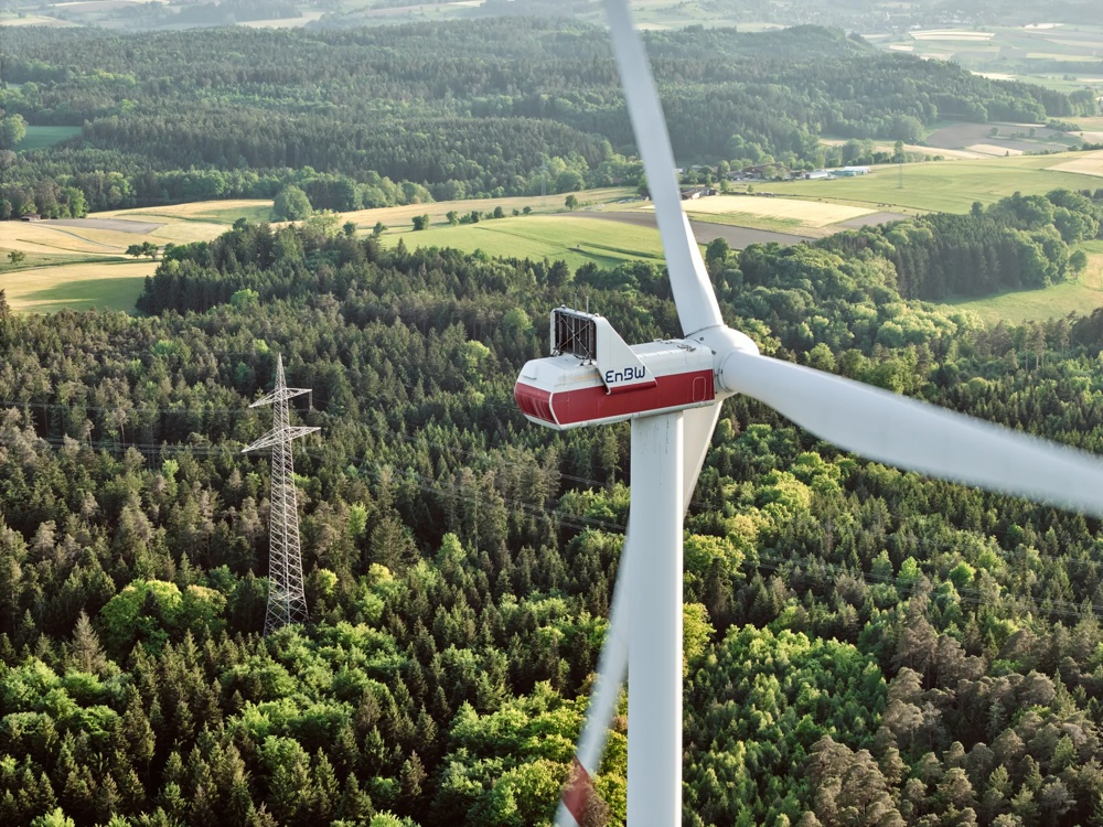 Drei Windräder stehen in einem grünen Wald unter blauem Himmel.