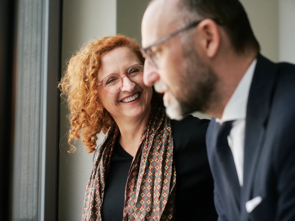 Eine lächelnde Frau mit lockigem Haar spricht mit einem Mann im Anzug vor einem Fenster.