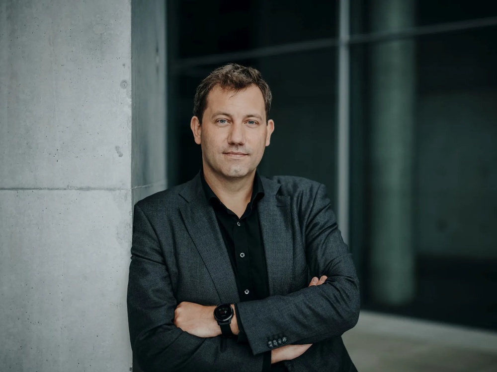 Professionelles Porträtfoto eines Mannes mittleren Alters mit kurzen braunen Haaren und blauen Augen. Er lehnt mit verschränkten Armen an einer Betonsäule, trägt einen dunkelgrauen Blazer über einem schwarzen Hemd und eine dunkle Smartwatch.