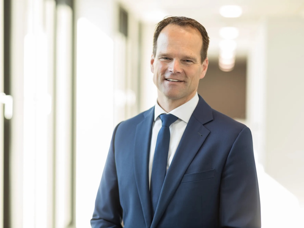 A man in a blue suit standing in a bright corridor smiling into the camera.