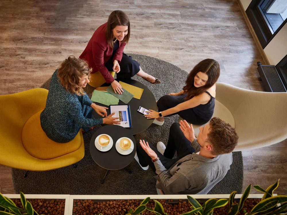Four people sitting around a round table with laptops, fabric samples and two cappuccinos on a yellow and beige armchair.