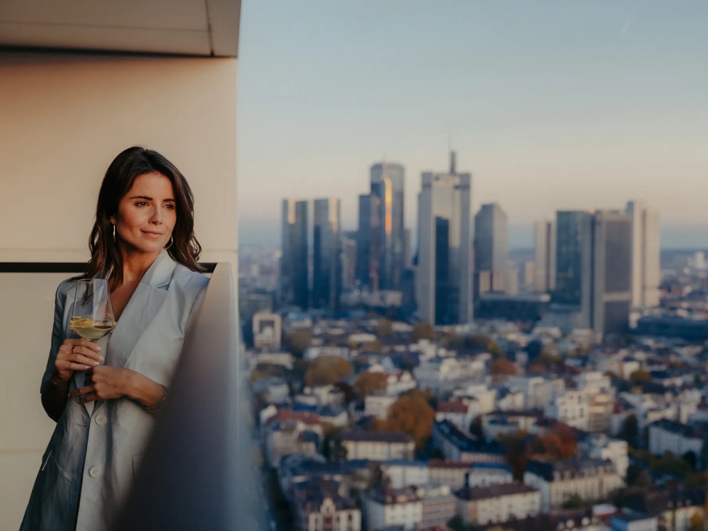 Person auf Balkon einer modernen Wohnung mit Blick über die Stadt und Getränk in der Hand.