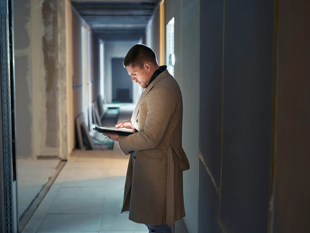 Ein Mann im Mantel steht in einem Flur und tippt auf ein Tablet.