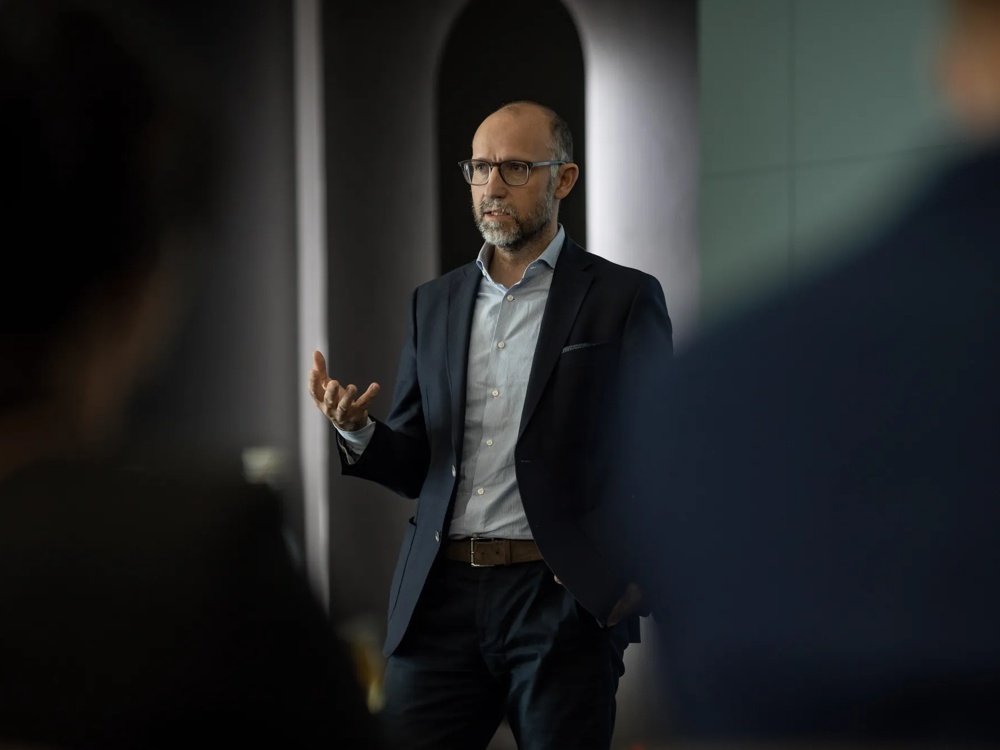 Ein Mann in Anzug spricht gestikulierend vor einem unscharfen Hintergrund.