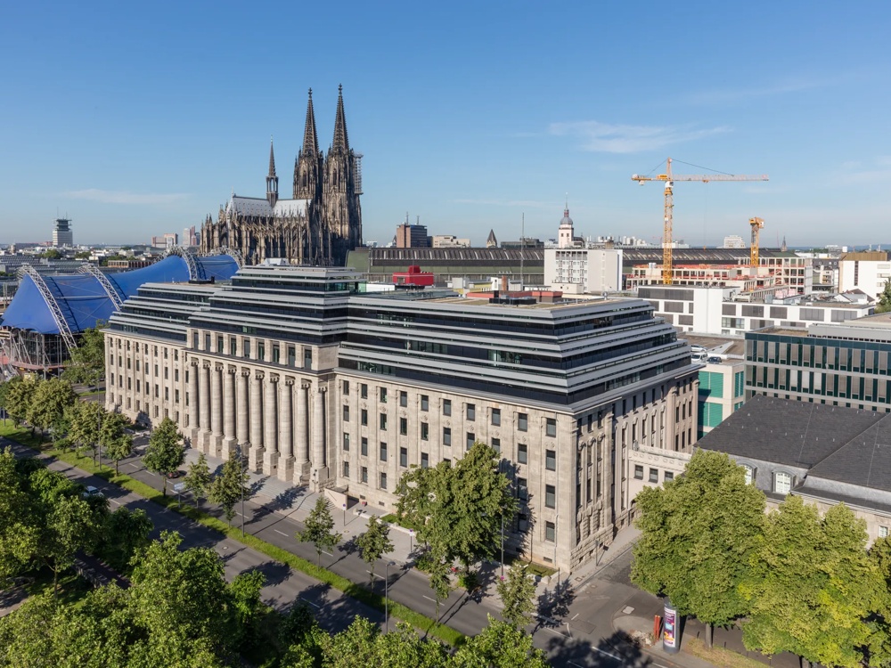 Historische Gebäude mit Kölner Dom im Hintergrund, blauer Himmel, Bäume und Baukräne.