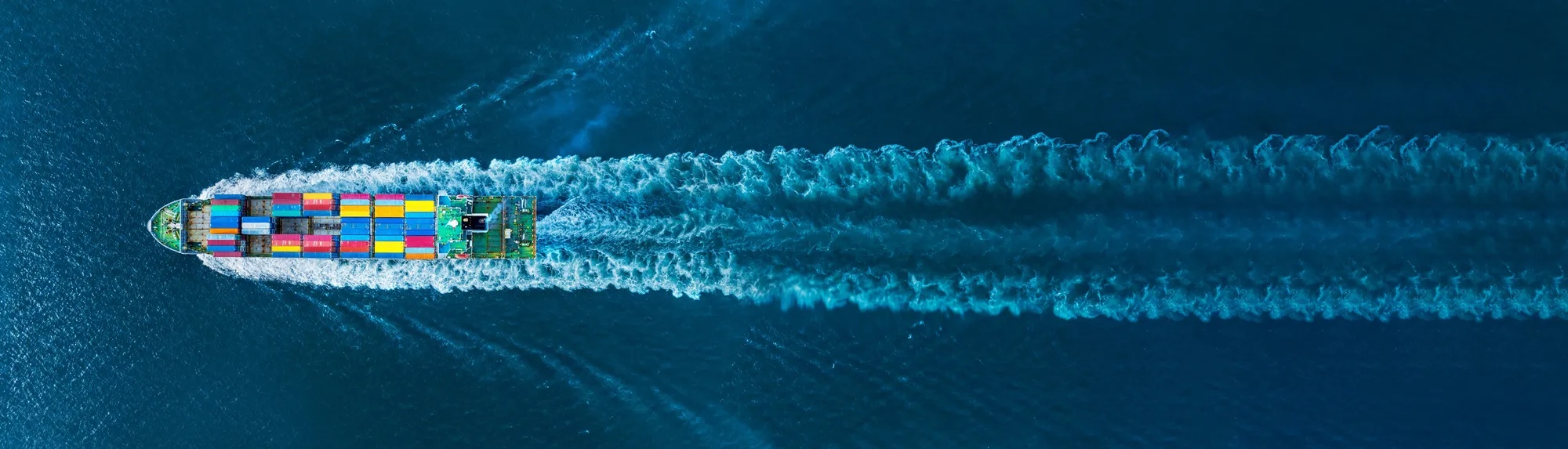Luftaufnahme eines Containerschiffs mit bunten Frachtcontainern, das auf tiefblauem Meer fährt und eine breite Bugwelle hinterlässt.
