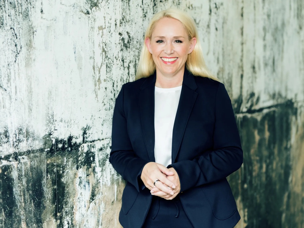 Frau in dunklem Anzug lächelt vor einer strukturierten, verwitterten Betonwand.